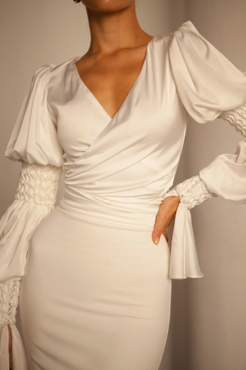 May include: A white satin dress with a V-neckline and long sleeves. The sleeves have a gathered detail at the cuffs. The dress is fitted and has a wrap-style bodice.