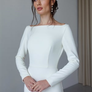 May include: A white long-sleeved wedding dress with a simple, elegant design. The dress has a bateau neckline and a fitted silhouette.