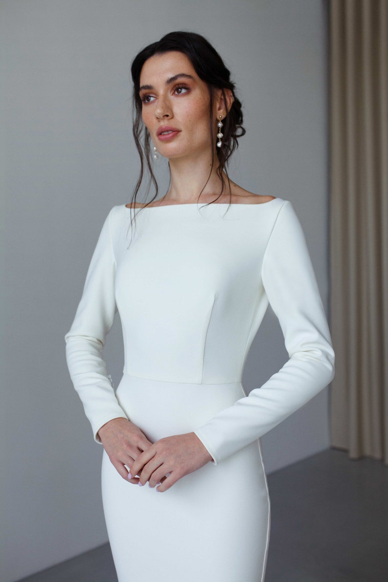 May include: A white long-sleeved wedding dress with a simple, elegant design. The dress has a bateau neckline and a fitted silhouette.