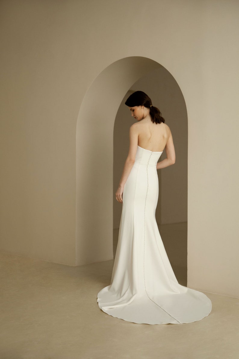 Puede incluir: Un vestido de novia blanco sin tirantes con un corpi&ntilde;o ajustado y una falda fluida. El vestido tiene una cola larga y est&aacute; hecho de una tela lisa y satinada.
