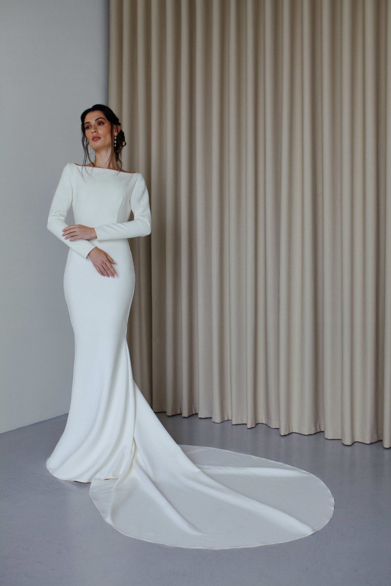 May include: A white long-sleeved wedding dress with a fitted bodice and a flowing train. The dress is made of a smooth, satin-like fabric.