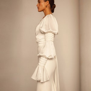 May include: A white satin dress with a fitted skirt and a fitted bodice. The dress has long sleeves with a gathered detail at the elbow and a ruffle at the wrist. The dress has a low neckline and a long train.