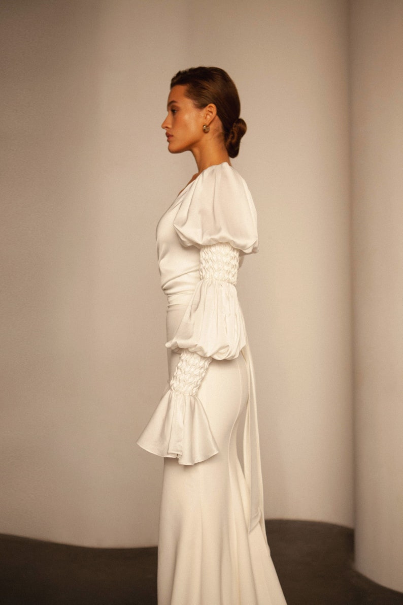 May include: A white satin dress with a fitted skirt and a fitted bodice. The dress has long sleeves with a gathered detail at the elbow and a ruffle at the wrist. The dress has a low neckline and a long train.