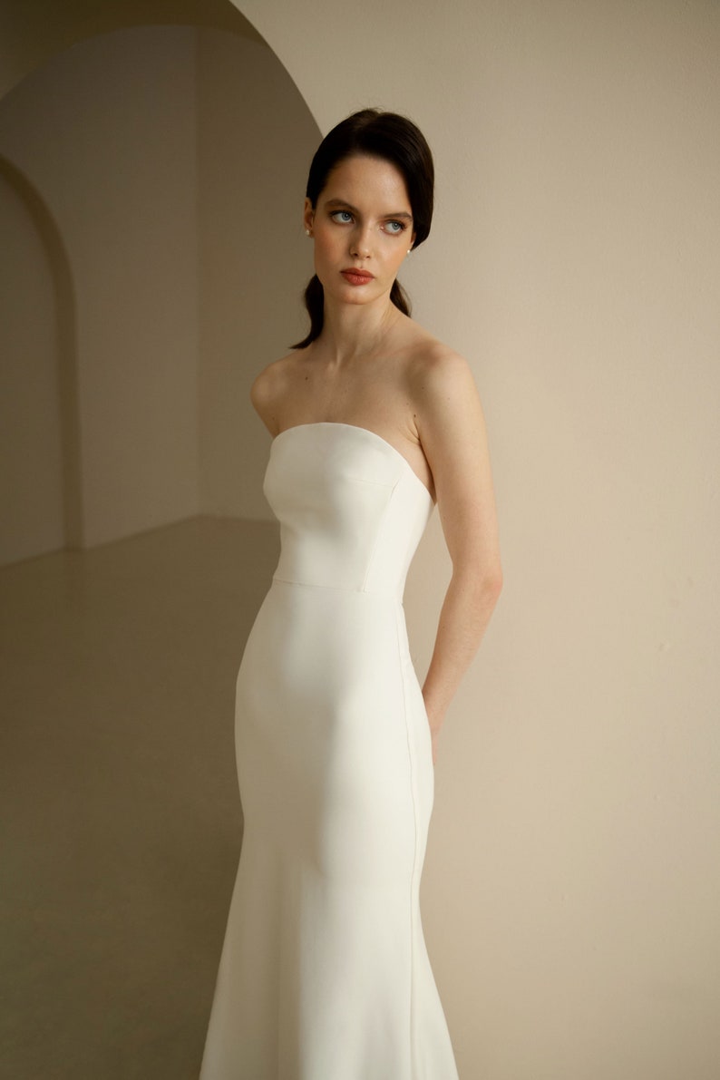 Puede incluir: Una mujer con un vestido de novia blanco sencillo y sin tirantes, con un corpi&ntilde;o ajustado y una falda fluida.