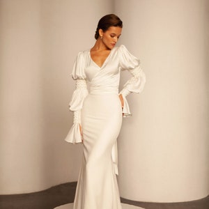 May include: A white satin wedding dress with a V-neckline, long sleeves, and a fitted silhouette. The dress features a wrap-style bodice and a flowing skirt.