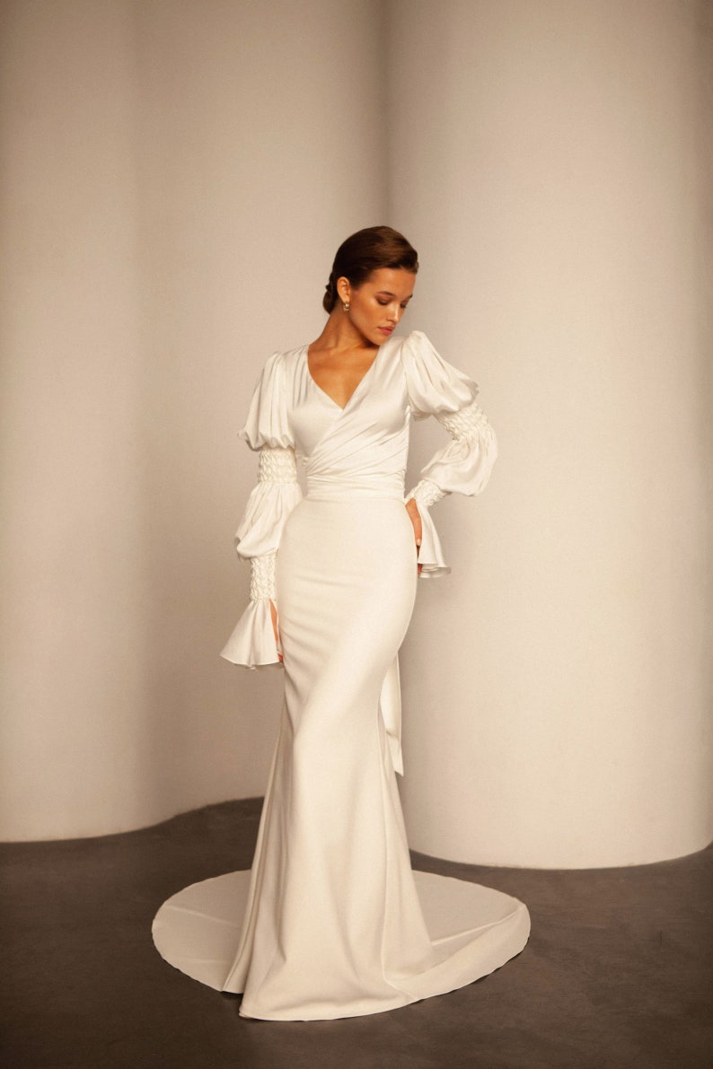 May include: A white satin wedding dress with a V-neckline, long sleeves, and a fitted silhouette. The dress features a wrap-style bodice and a flowing skirt.