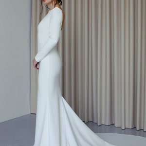 May include: A white long-sleeved wedding dress with a fitted bodice and a flowing skirt. The dress has a low back and a train.