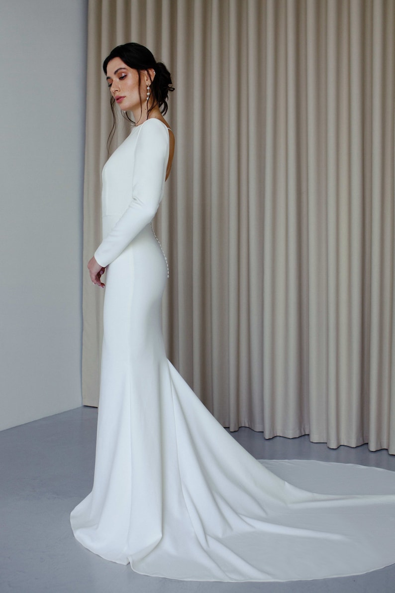May include: A white long-sleeved wedding dress with a fitted bodice and a flowing skirt. The dress has a low back and a train.