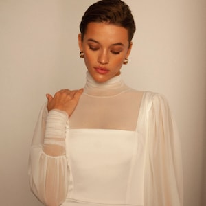 May include: A white dress with a high neck and long sleeves. The sleeves are sheer and have a gathered detail at the shoulder. The dress is fitted through the bodice and has a flowing skirt.
