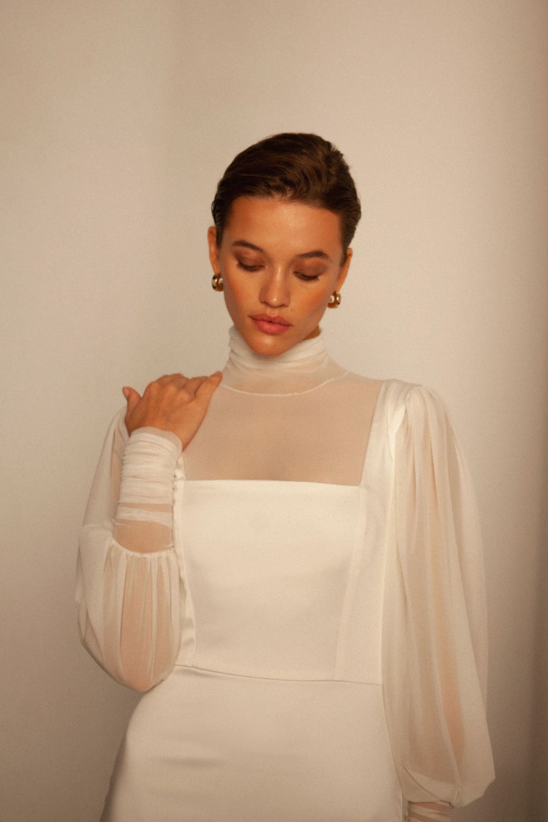 May include: A white dress with a high neck and long sleeves. The sleeves are sheer and have a gathered detail at the shoulder. The dress is fitted through the bodice and has a flowing skirt.