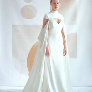 Puede incluir: Un vestido de novia blanco con cuello alto y una larga cola. El vestido tiene un corpi&ntilde;o ajustado y una falda fluida. La modelo est&aacute; de pie frente a un fondo blanco con formas abstractas.