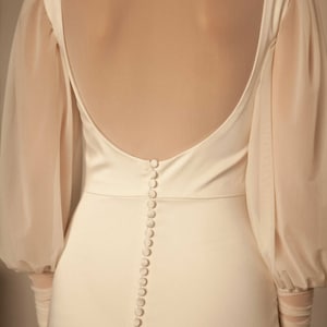 May include: A white satin dress with a low back and a row of buttons down the centre. The dress has long sleeves with sheer fabric at the cuffs.