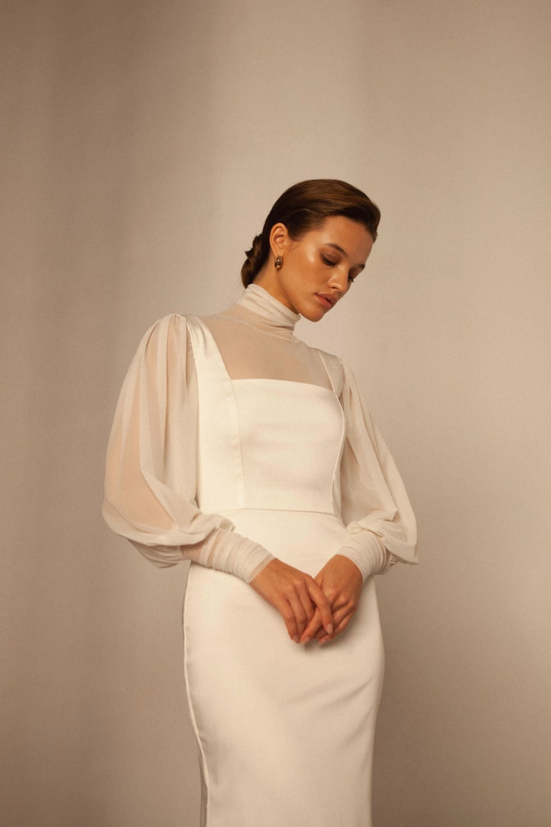 May include: A white dress with a high neck and long sleeves. The sleeves are sheer and have a puffed design. The dress has a fitted bodice and a skirt that falls to the knees.