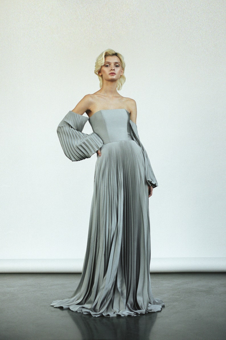 May include: A silver off-the-shoulder gown with a pleated skirt and large, flowing sleeves.