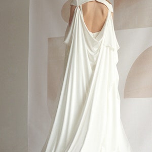 Puede incluir: Un vestido de novia blanco con una larga cola y una espalda dram&aacute;tica en forma de capa. El vestido tiene un escote alto y una espalda baja, con recortes en los hombros.