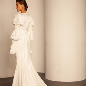 May include: A white wedding dress with a fitted bodice and a long train. The dress has long sleeves with puffed shoulders and a satin sash.
