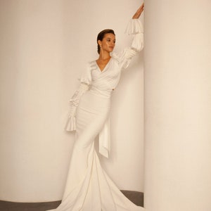 May include: A white, long-sleeved, wrap-style dress with a fitted bodice and a flowing skirt. The dress has a V-neckline and ruched sleeves.