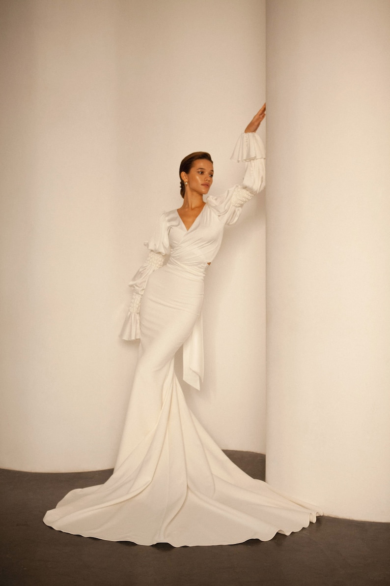May include: A white, long-sleeved, wrap-style dress with a fitted bodice and a flowing skirt. The dress has a V-neckline and ruched sleeves.