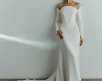 May include: A white, off-the-shoulder wedding dress with long sleeves and a flowing train. The gown has a fitted bodice and a streamlined silhouette. The model is standing against a white wall, highlighting the elegant design of the bridal gown.