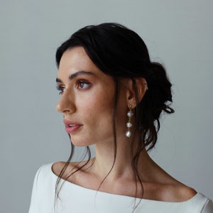May include: A woman with dark hair wearing a white off-the-shoulder dress and gold earrings with pearl accents.