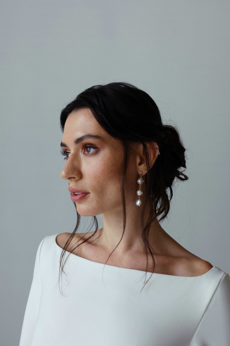 May include: A woman with dark hair wearing a white off-the-shoulder dress and gold earrings with pearl accents.
