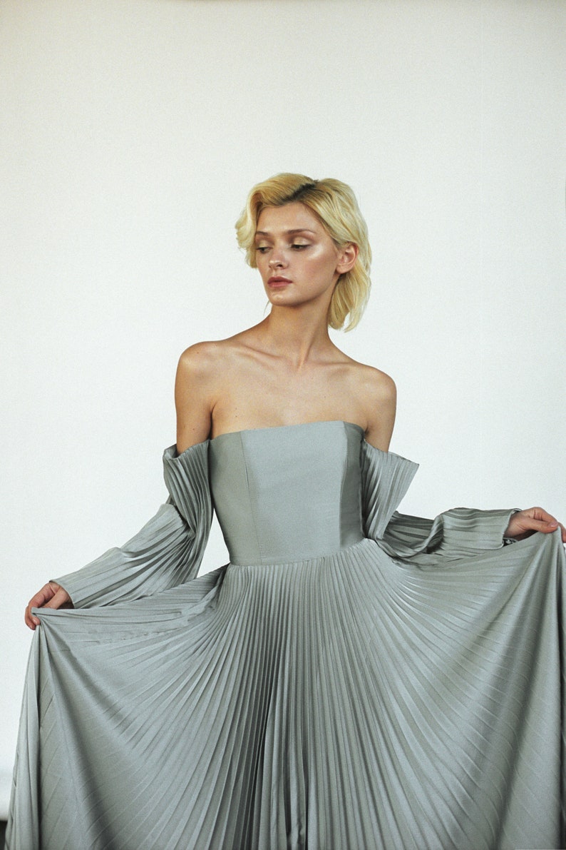 May include: A woman wearing a silver off-the-shoulder gown with pleated fabric. The gown has long sleeves and a fitted bodice.