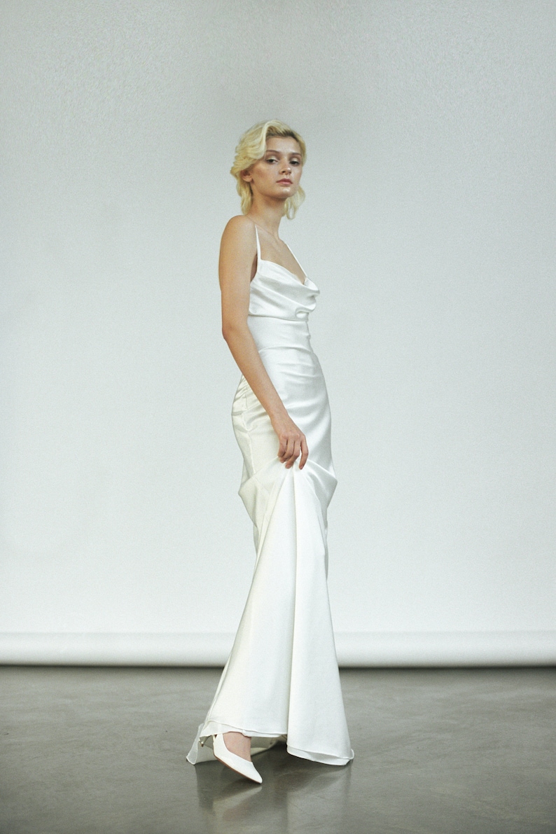 Peut inclure: Une femme portant une robe blanche en satin &agrave; col en V avec une longue tra&icirc;ne. Elle est debout sur un sol gris et regarde la cam&eacute;ra.