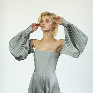 May include: A woman wearing a silver off-the-shoulder dress with large, pleated sleeves. The dress has a fitted bodice and a flowing skirt.