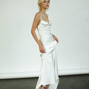 Peut inclure: Une femme portant une robe blanche en satin avec un col en V et une jupe fluide. Elle se tient debout sur une surface noire et tient la jupe de sa main droite.