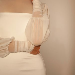 May include: A white dress with a high neck and long sleeves. The sleeves are sheer and have a gathered detail at the shoulders.