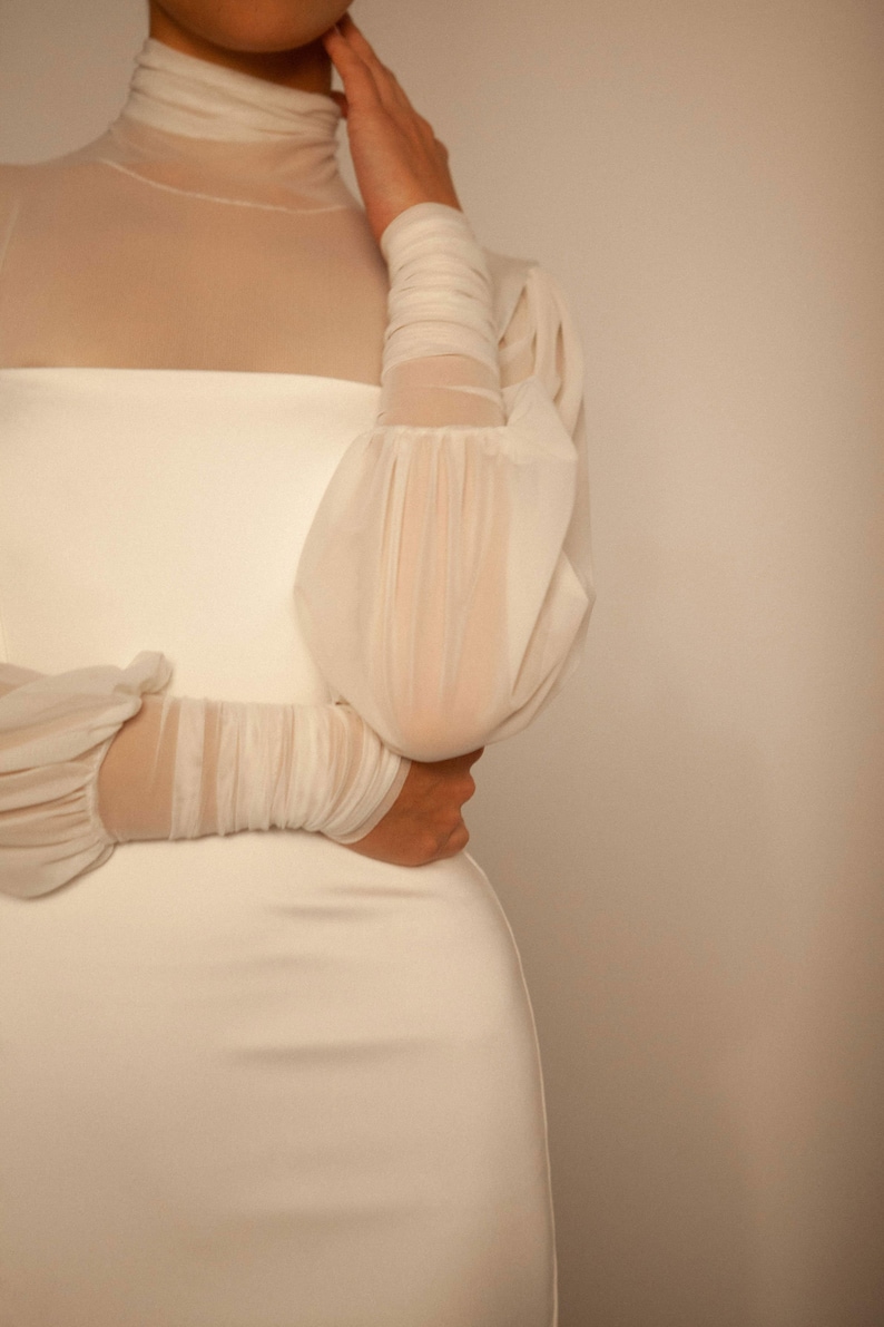 May include: A white dress with a high neck and long sleeves. The sleeves are sheer and have a gathered detail at the shoulders.