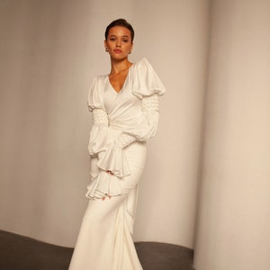 May include: A white satin wedding dress with a fitted bodice and a flowing skirt. The dress features long sleeves with puffed shoulders and ruffles at the cuffs.