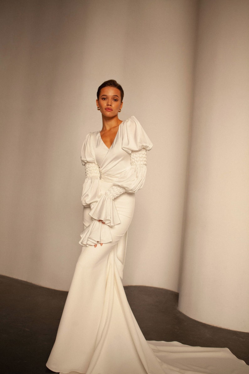 May include: A white satin wedding dress with a fitted bodice and a flowing skirt. The dress features long sleeves with puffed shoulders and ruffles at the cuffs.
