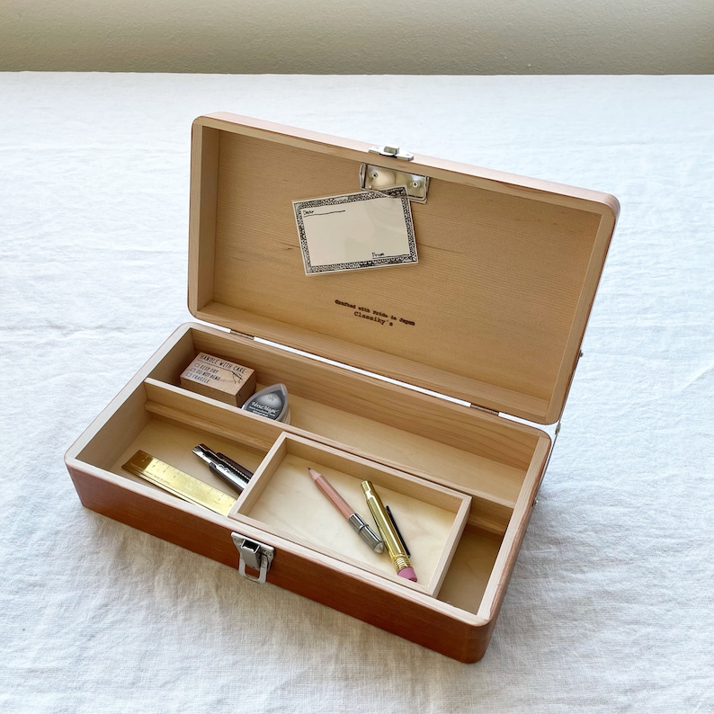 May include: A wooden box with a hinged lid, open to reveal a variety of small items, including a ruler, pencils, a stamp, and a pencil sharpener. The box is made of light wood and has a natural finish. The text " Crafted with Pride in Japan Cleverly." is printed on the inside of the lid.