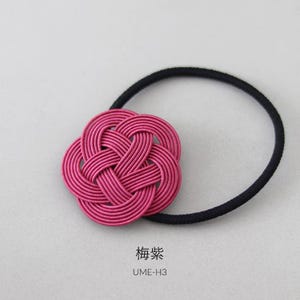 Puede incluir: Una liga para el pelo trenzada de color rosa con un nudo decorativo en forma de flor. La liga para el pelo est&aacute; sobre un fondo blanco. El texto "梅染" y "UME-H3" est&aacute; impreso debajo de la liga para el pelo.