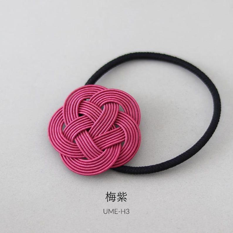 Puede incluir: Una liga para el pelo trenzada de color rosa con un nudo decorativo en forma de flor. La liga para el pelo est&aacute; sobre un fondo blanco. El texto "梅染" y "UME-H3" est&aacute; impreso debajo de la liga para el pelo.