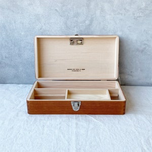 May include: A wooden box with a hinged lid and a metal latch. The box has a compartment with dividers inside. The box is made of light brown wood and has a dark brown stain. The box has the text "crafted with pride in Japan" and "Classicky" on the inside of the lid.