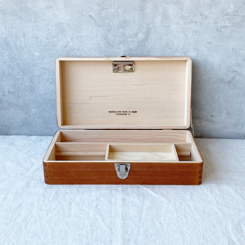 May include: A wooden box with a hinged lid and a metal latch. The box has a compartment with dividers inside. The box is made of light brown wood and has a dark brown stain. The box has the text "crafted with pride in Japan" and "Classicky" on the inside of the lid.
