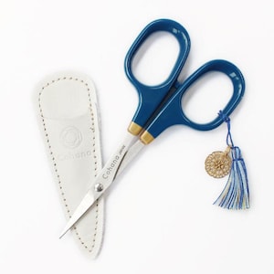 May include: Dark blue scissors with gold-toned accents at the pivot point and a silver blade engraved with the text Cohana JAPAN. The scissors feature a blue and white tassel with a gold-toned circular charm and a white leather protective sheath.