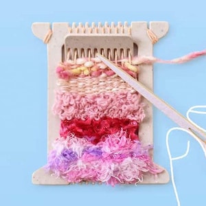 May include: A white wooden loom with pink, red, and purple yarn woven into a tapestry. A hand is holding a wooden weaving tool and guiding the yarn.