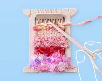 Peut inclure: Un métier à tisser en bois blanc avec du fil rose, rouge et violet tissé en tapisserie. Une main tient un outil de tissage en bois et guide le fil.