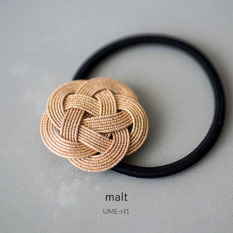 Puede incluir: Una liga para el pelo tejida en beige con una banda el&aacute;stica negra. La liga para el pelo tiene forma de c&iacute;rculo con un dise&ntilde;o de nudo. El texto "malt UME-H1" est&aacute; impreso debajo de la liga para el pelo.