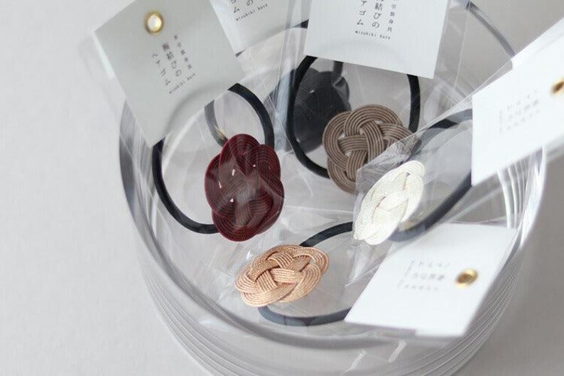 Puede incluir: Primer plano de cuatro gomas para el pelo con nudos de diferentes colores. Las gomas para el pelo est&aacute;n en un recipiente transparente con etiquetas blancas que tienen texto japon&eacute;s.