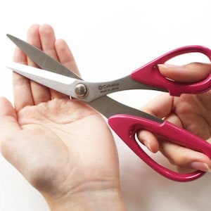Könnte beinhalten: Silberne Schere mit leuchtend pinkfarbenen Griffen. Die Klingen sind geöffnet und zeigen die scharfen Kanten und den Markennamen "Cohana". Diese Schere ist für Bastelarbeiten und Nähen konzipiert. Die Griffe sind in einer hellen, auffälligen Farbe gehalten.