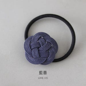 Puede incluir: Una liga para el pelo de color morado con un nudo decorativo en el centro. El nudo est&aacute; hecho de tela trenzada y tiene una apariencia texturizada. La liga para el pelo est&aacute; etiquetada con "梅黒" y "UME-H5".