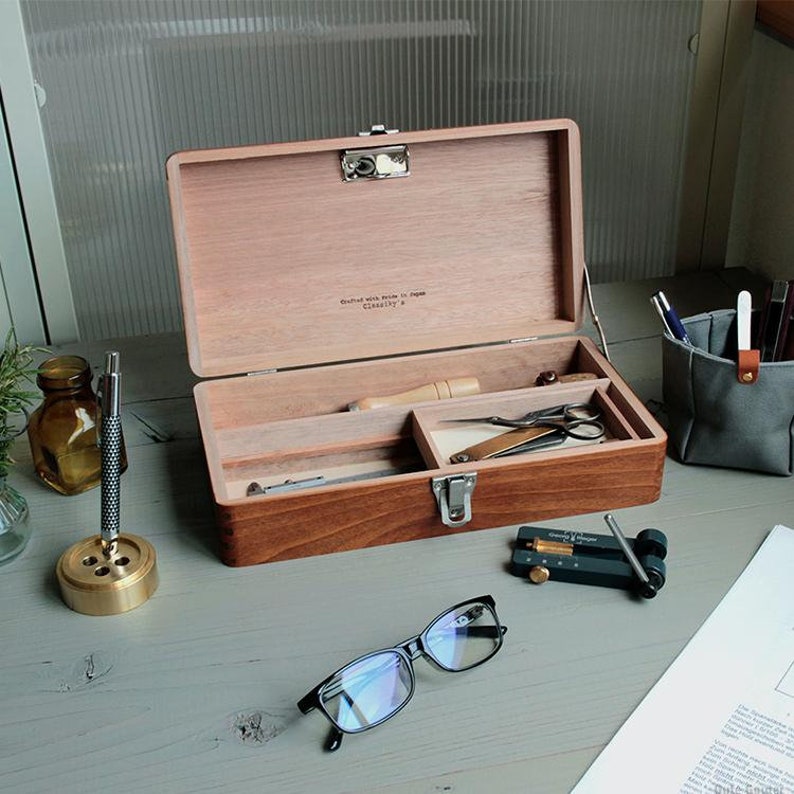 May include: A wooden box with a hinged lid and a metal latch. The box is divided into three compartments and contains various tools, including a pair of scissors, a small knife, and a needle. The box is labeled "crafted with pride in Japan" and "quality &"