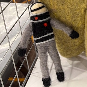 May include: A handmade felt robot doll with a black torso, gray limbs, and a white helmet. The robot has a red heart-shaped detail on its chest and is hanging against a yellow background. The doll is a unique, handcrafted item.