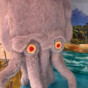 May include: A handmade, lavender-colored felted octopus sculpture. The octopus has large, round eyes with yellow and red details. Its tentacles are hanging down. The sculpture is displayed on a blue surface.