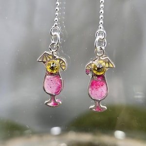 May include: A pair of silver-toned cocktail earrings. Each earring features a cocktail glass design with a pink drink, a yellow lemon slice, and a yellow gem. The earrings hang from a delicate chain.