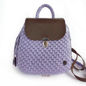 May include: A lavender crocheted backpack with a brown leather flap and a silver clasp. The backpack has a braided strap and tassels.